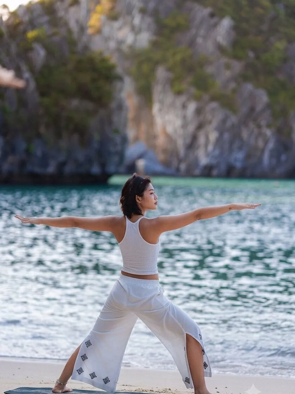 A person in white clothes practises yoga on a beach beside turquoise water and cliffs.