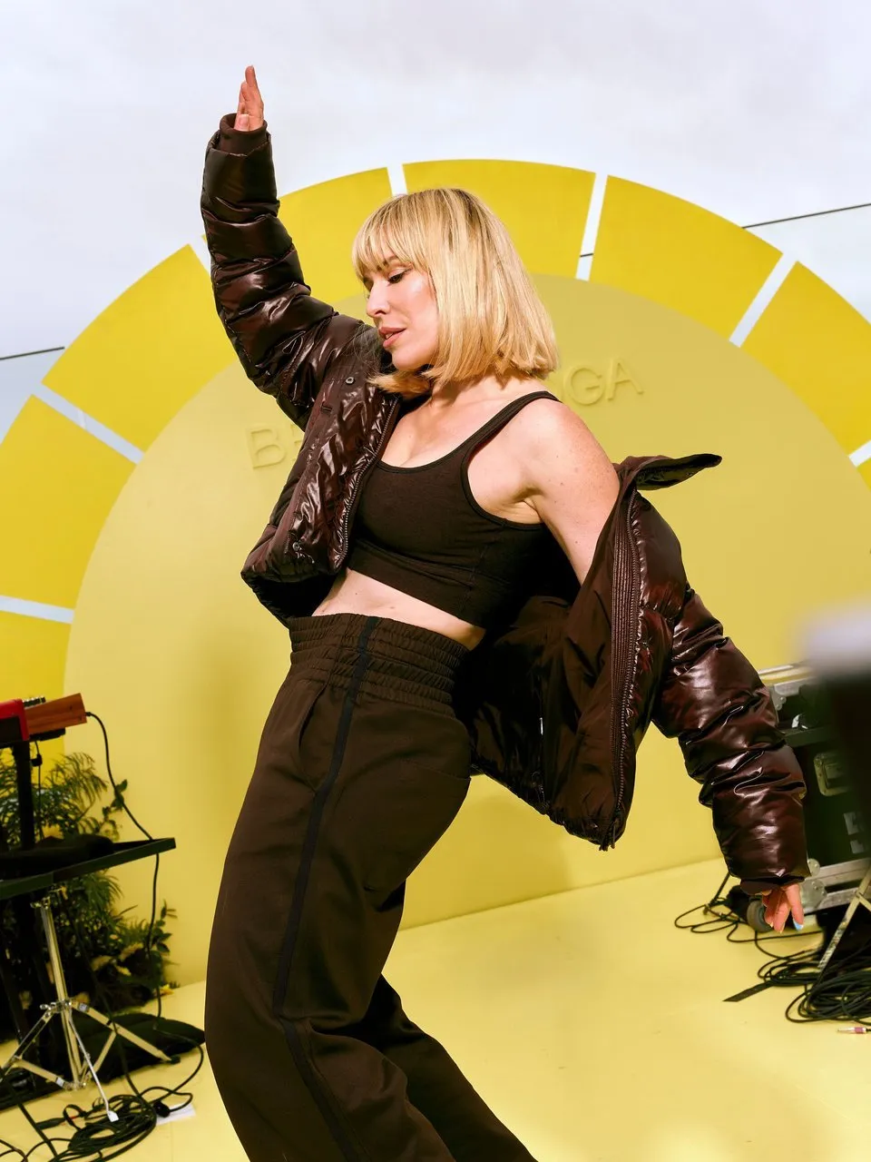 A person in a brown puffer jacket and black outfit poses in front of a bright yellow backdrop.