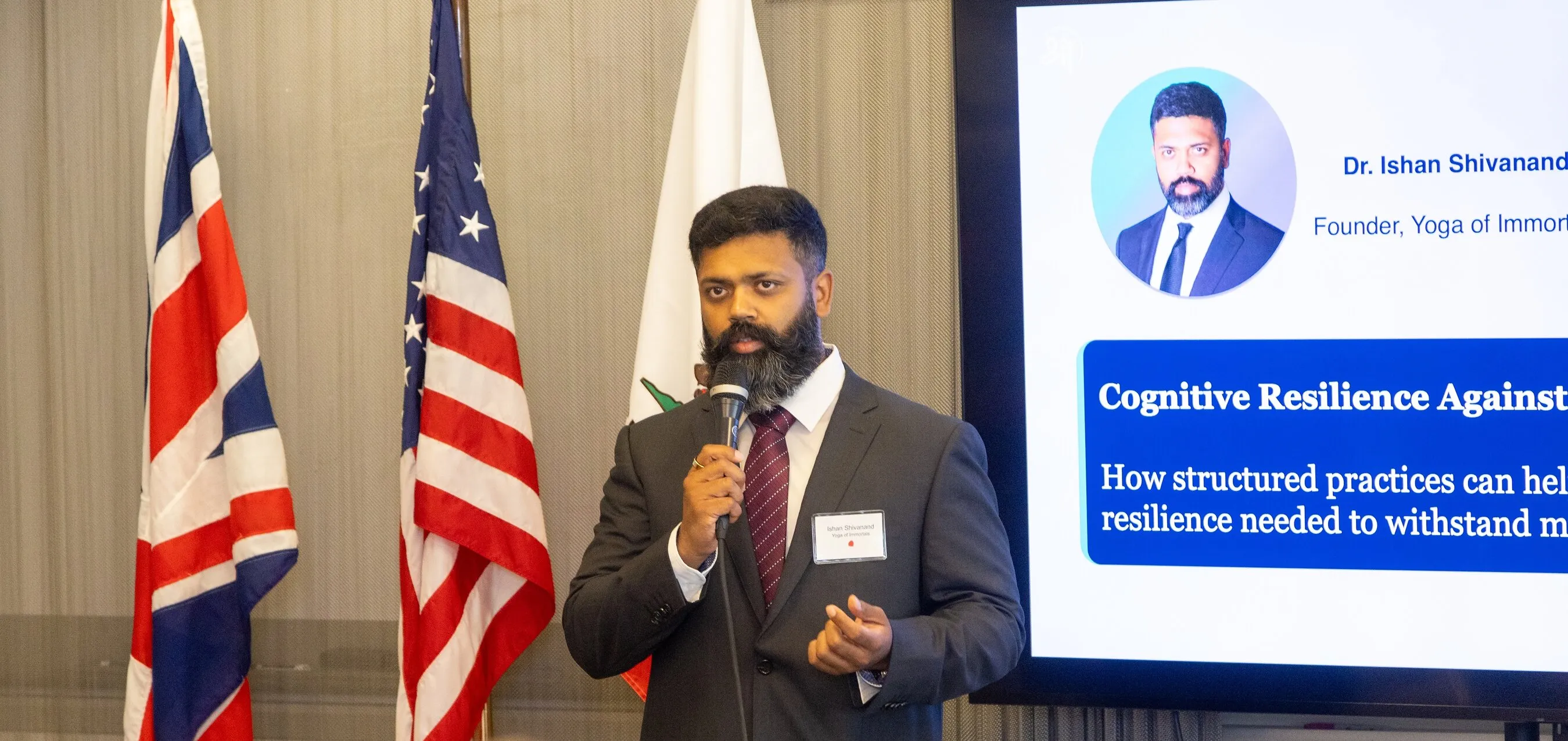 A person in a suit speaks into a microphone beside UK and US flags.