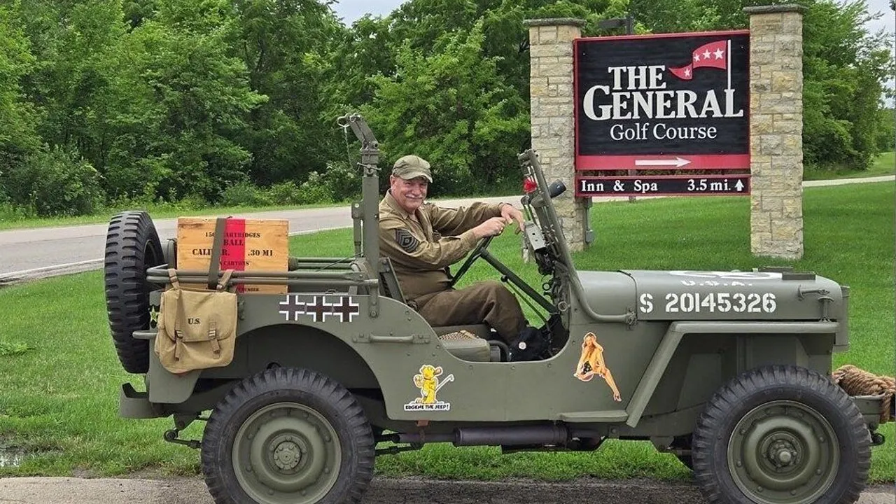 A person in military uniform drives a vintage jeep beside a golf course sign.