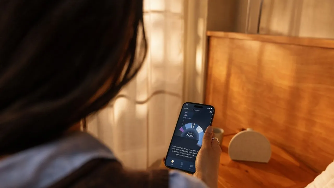 A person holds a phone showing a sleep-tracking app beside a bed in warm sunlight.