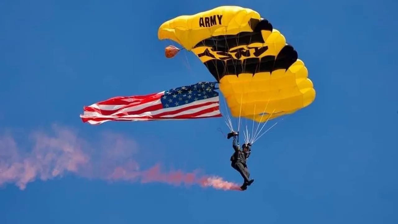 A U.S. Army parachutist descends beneath a yellow-and-black canopy carrying an American flag.