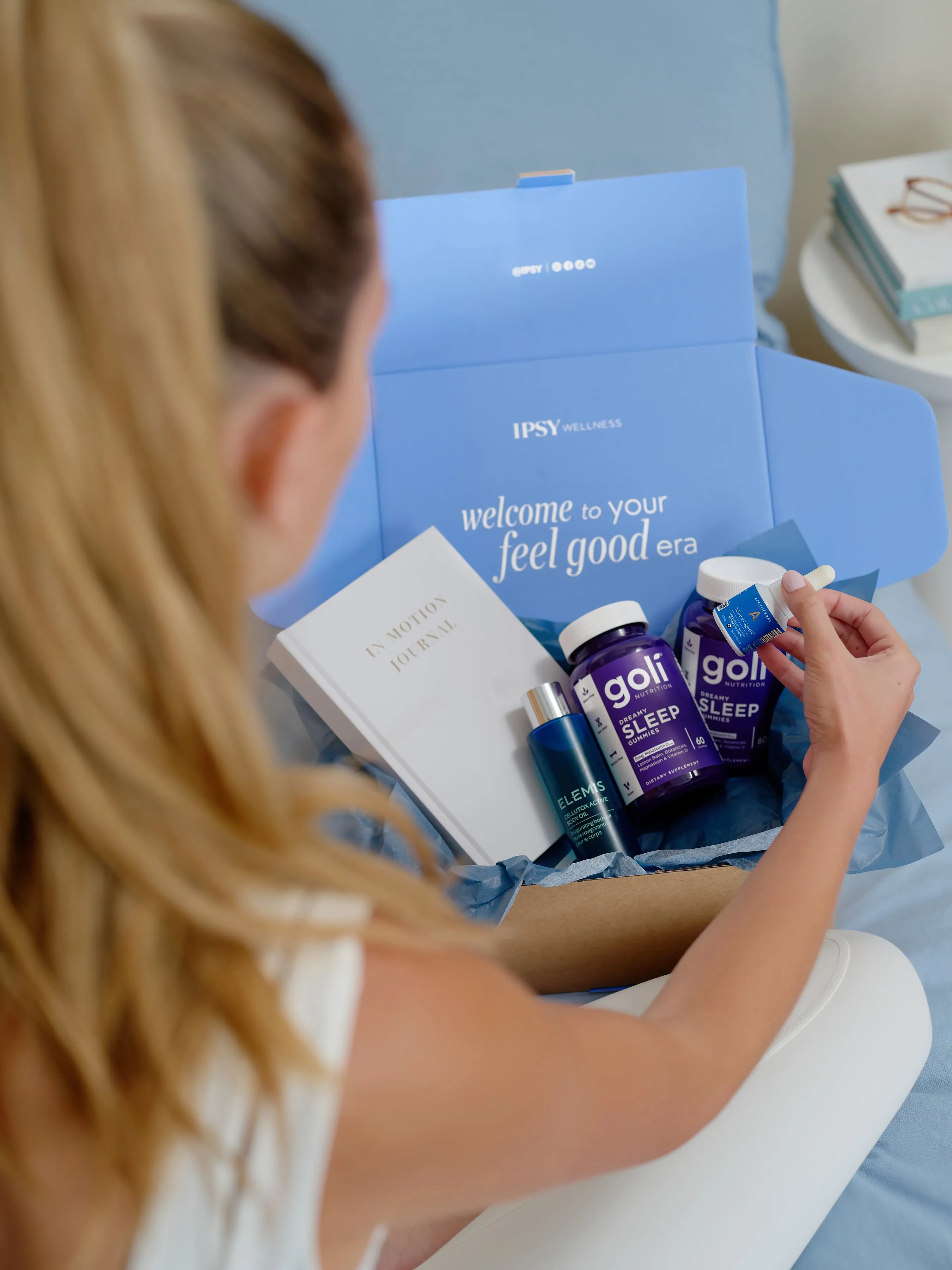 A person holds a supplement jar over an IPSY Wellness box filled with sleep gummies, skincare, and a journal.
