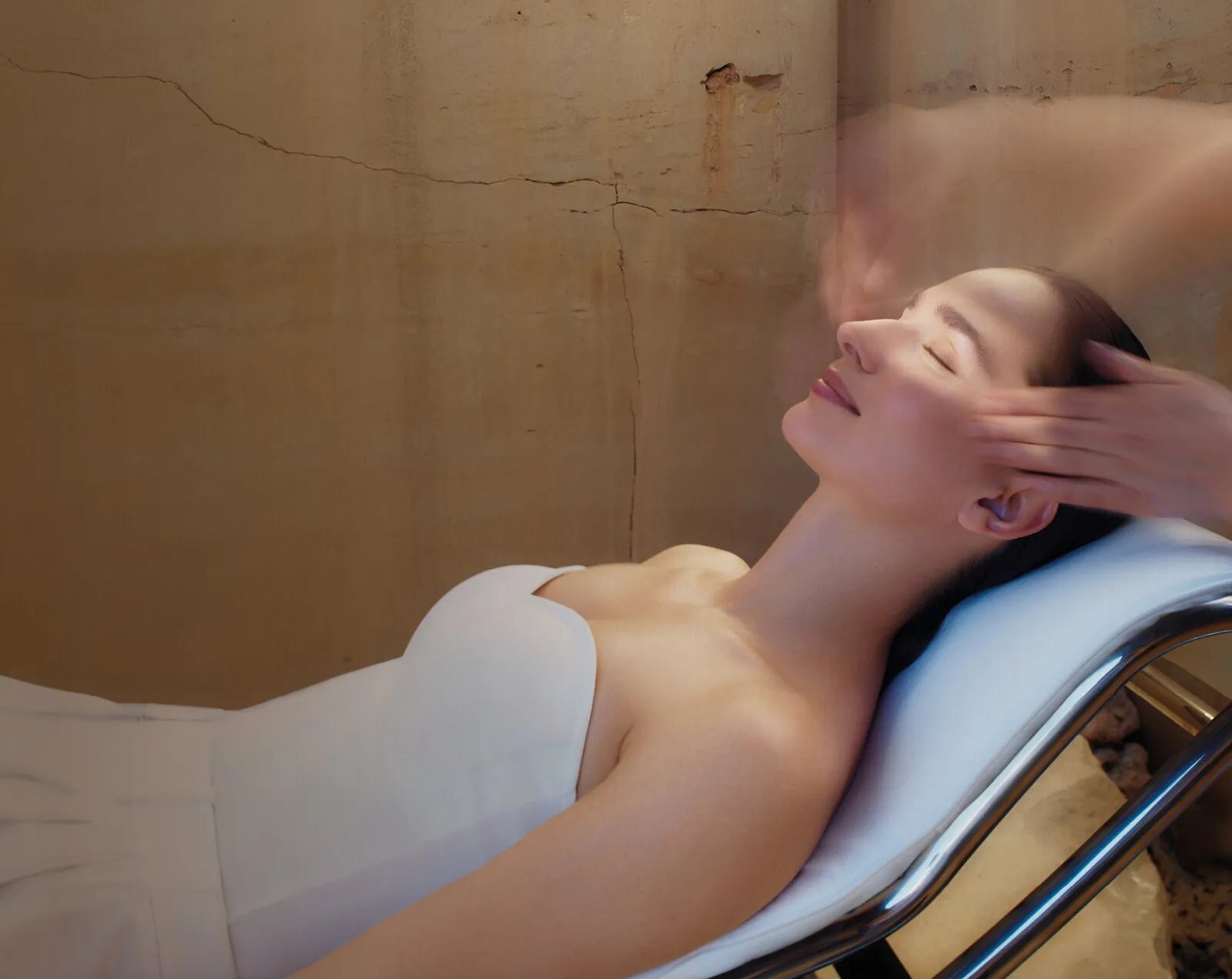 A person relaxes on a spa treatment bed while hands massage her temples.