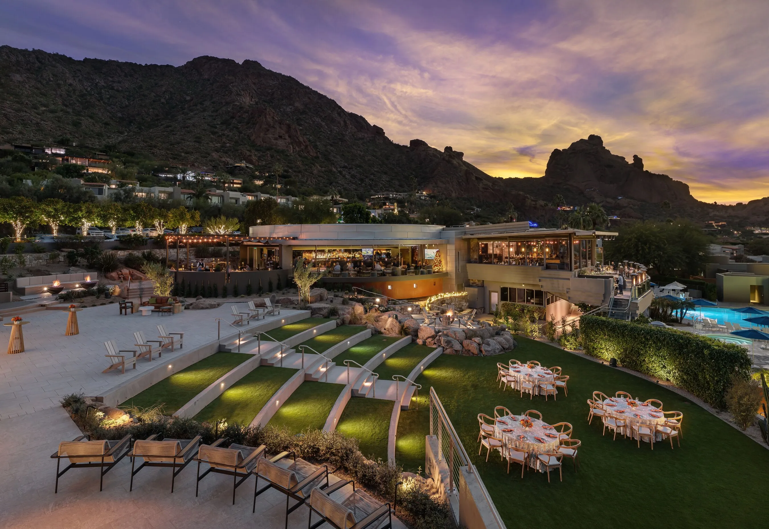 Terraced lawn seating and dining tables below a patio restaurant at sunset, framed by rocky mountains.