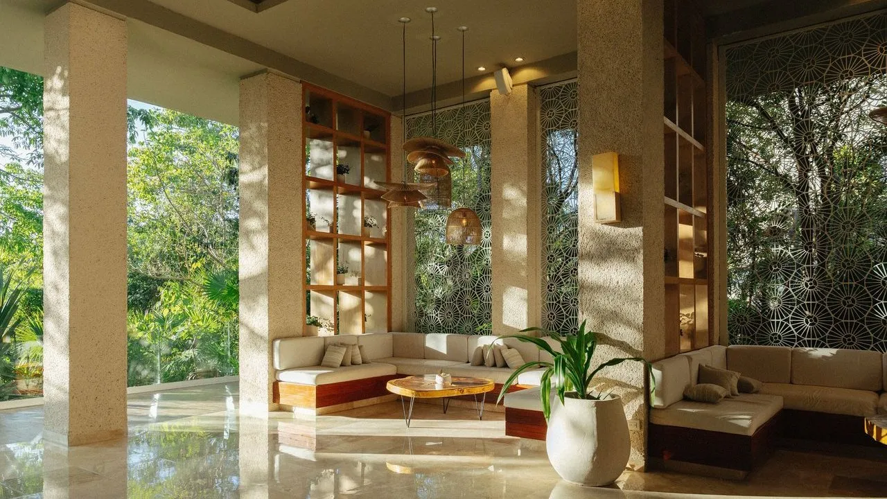 Sunlit tropical hotel lounge with sofas, hanging lights, patterned screens and lush garden views.