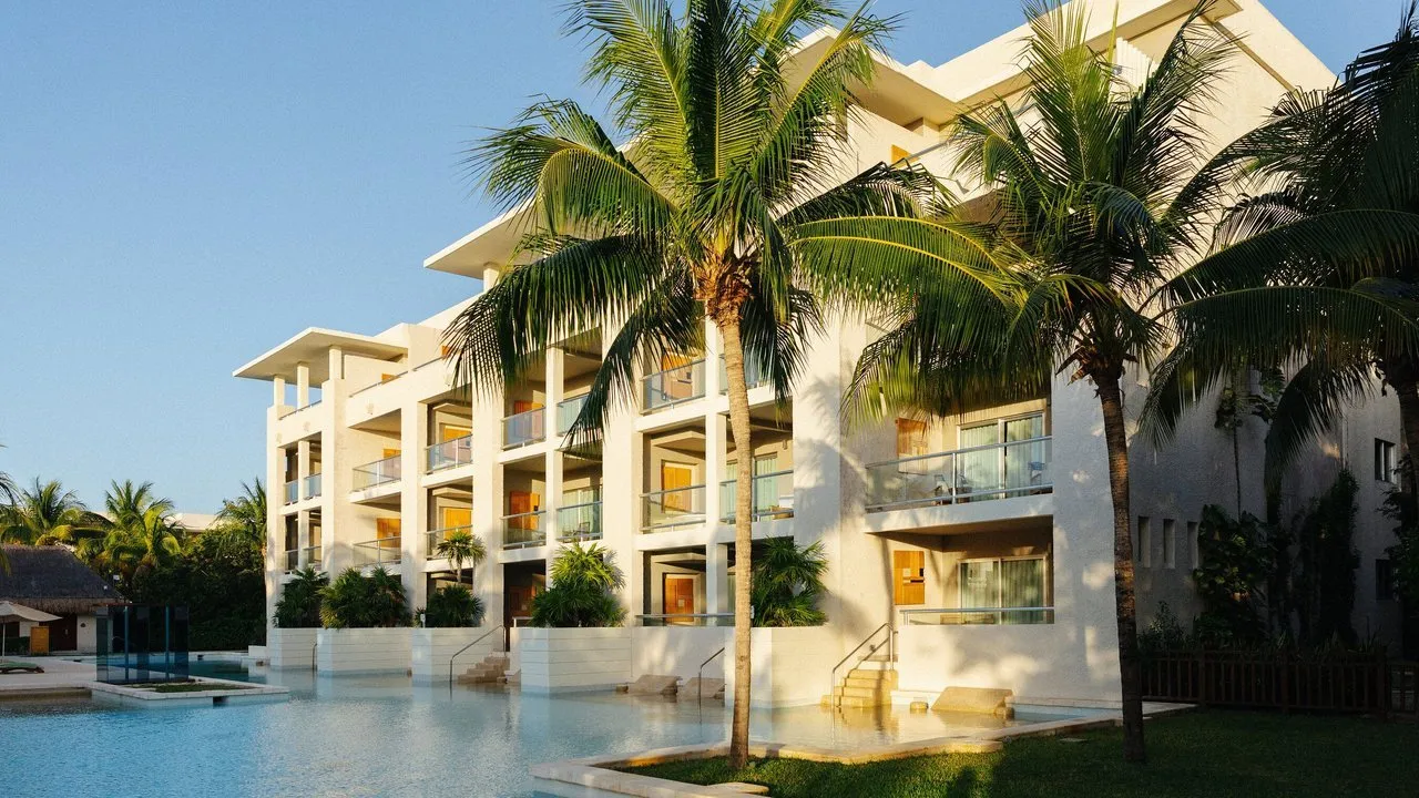 A white resort building with balconies, palm trees and a swimming pool.
