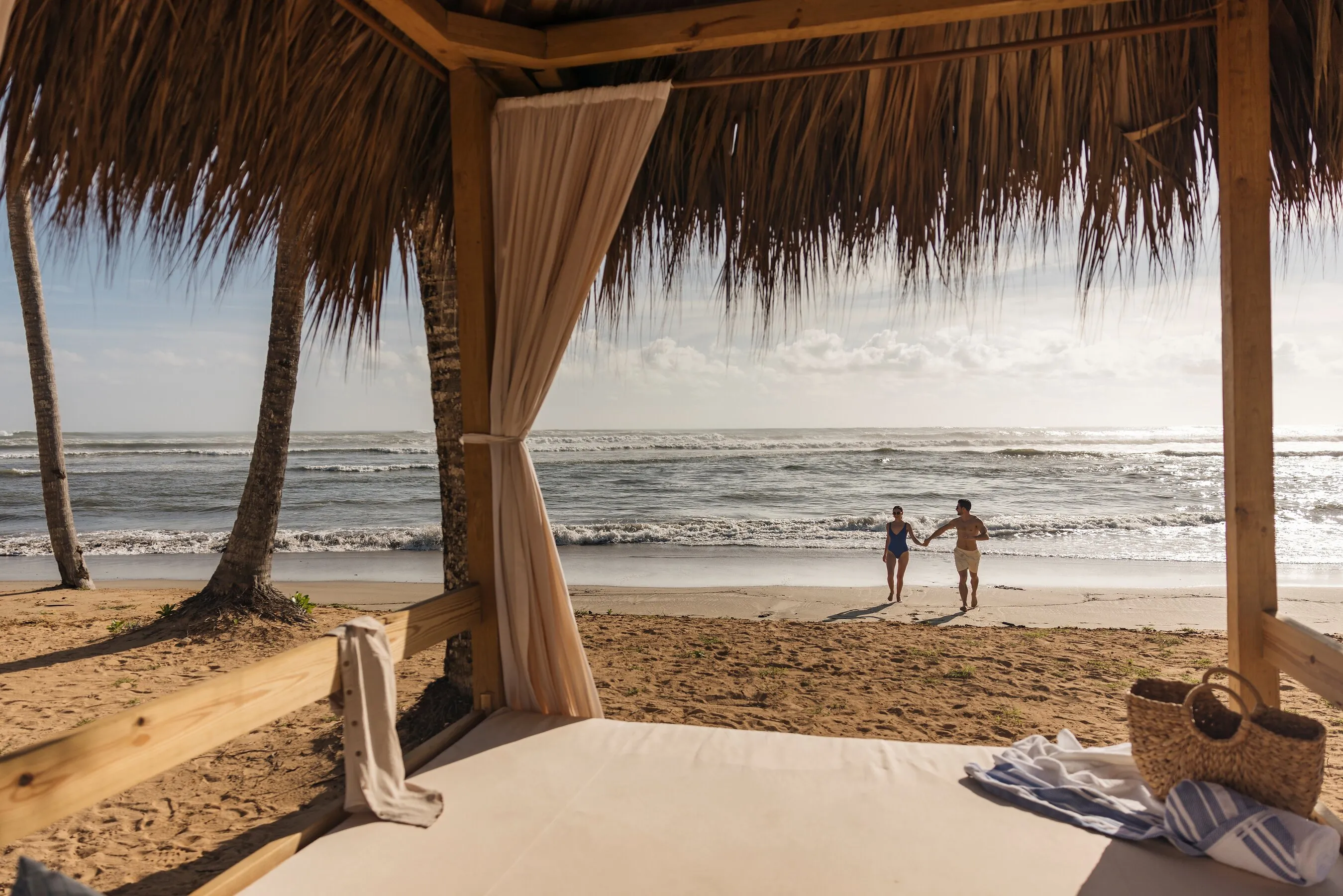 A beach cabana overlooks two people walking hand in hand beside the ocean.