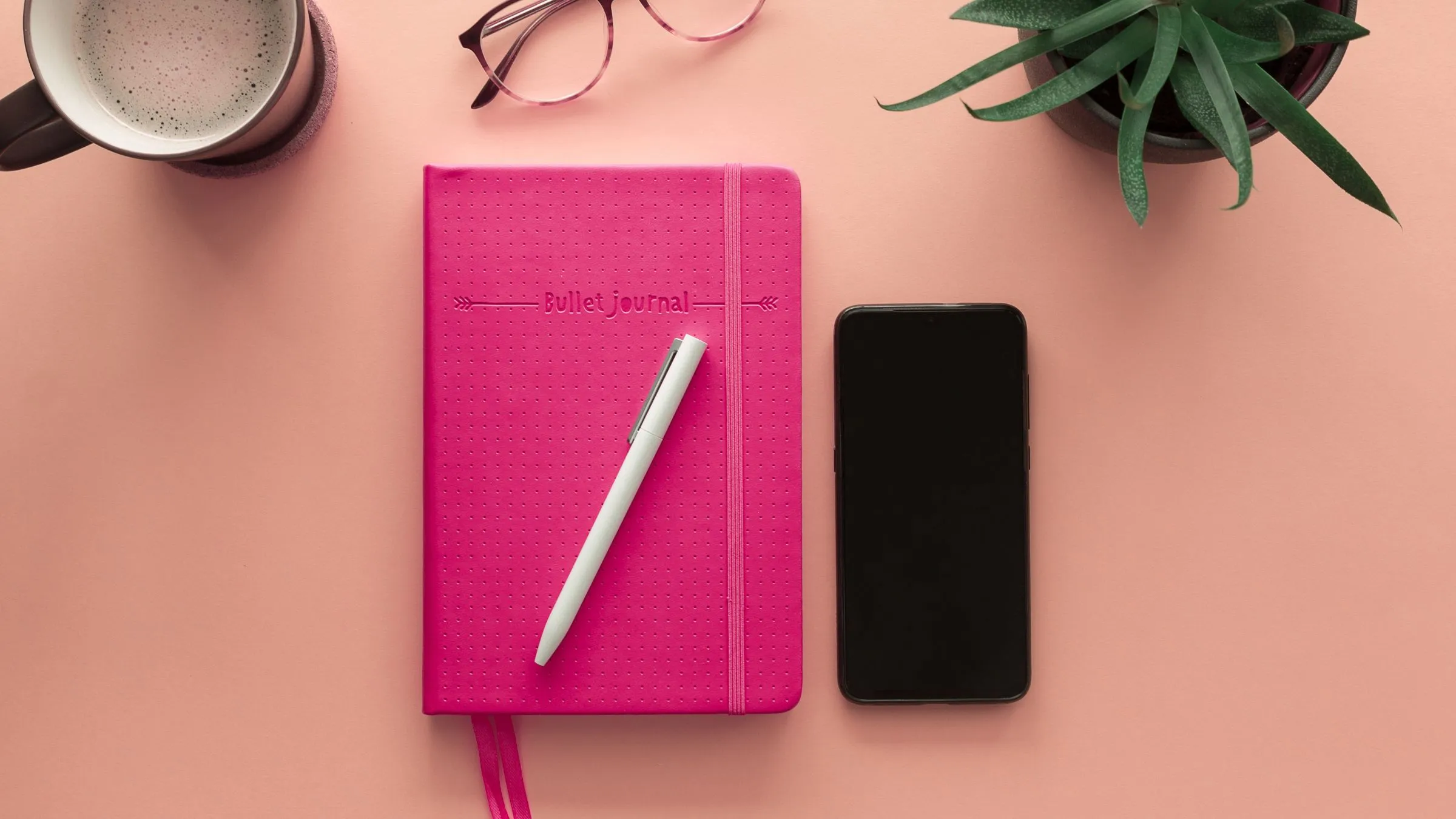 A pink journal and pen beside a phone, coffee and a plant on a pink desk.