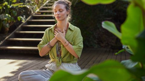 A woman meditating on a garden deck with her hands on her chest.