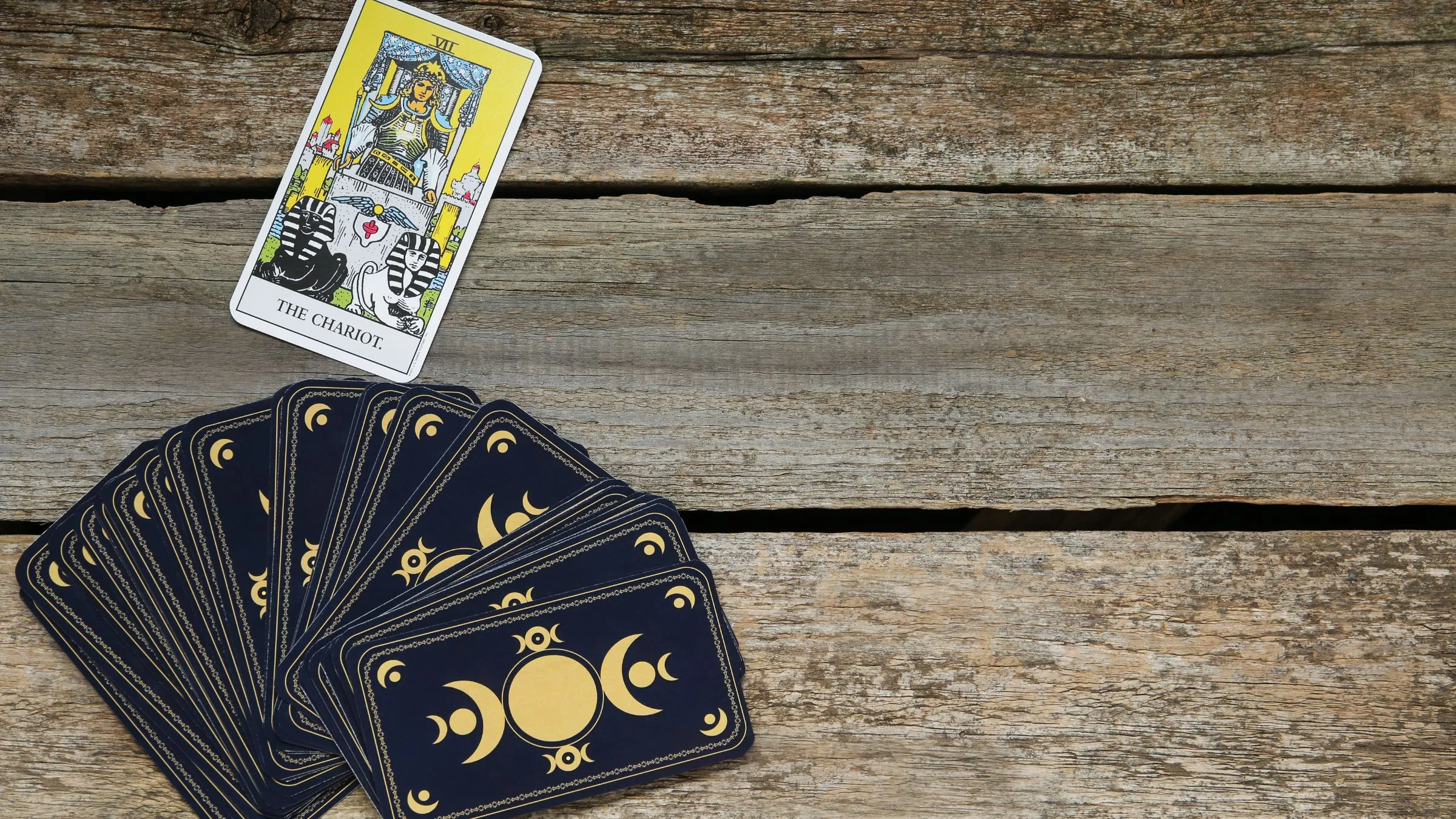 The Chariot tarot card above a fan of dark card backs on weathered wood.