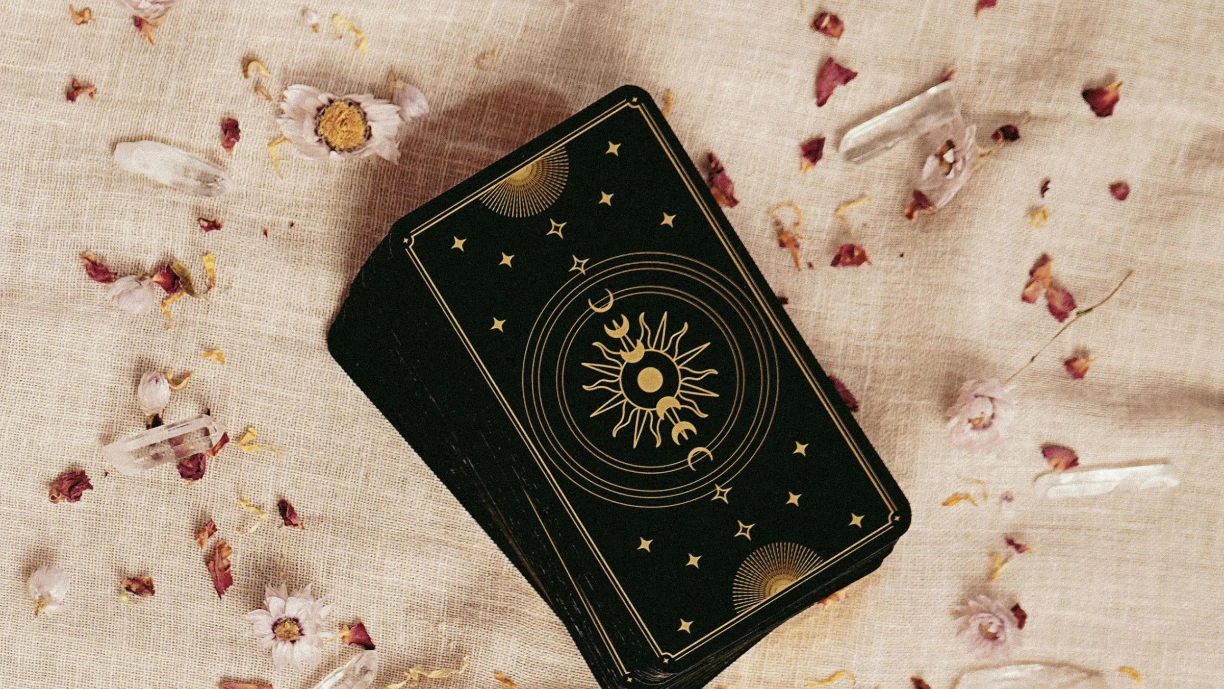 A black and gold tarot deck on linen scattered with dried petals and quartz.