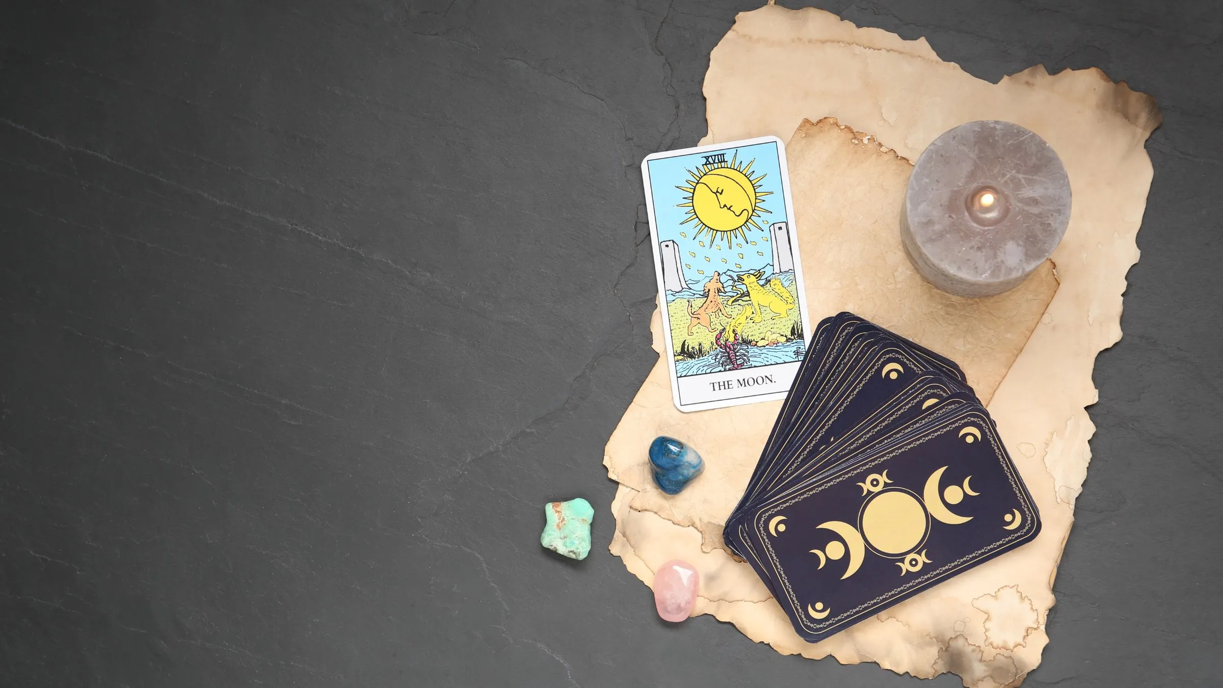 The Moon tarot card on old paper with a candle, crystals and card backs on slate.