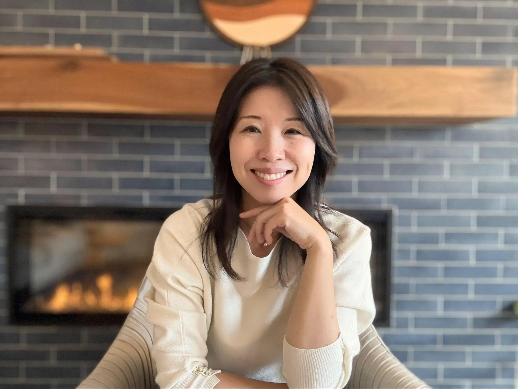 A smiling woman sits in front of a fireplace.