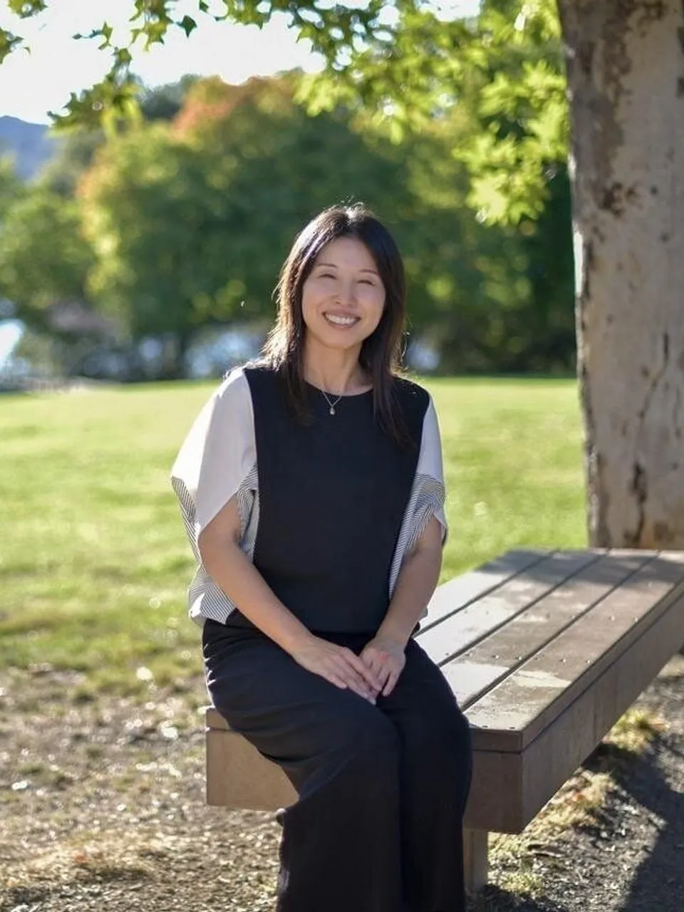 A smiling woman sits on a wooden bench in a sunny park beside a tree.